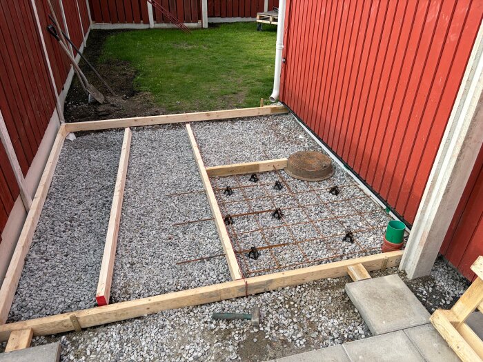 Betongform med armeringsjärn på grus, omgiven av röd husfasad. Form är 9x11 ft. Spade och grön gräsmatta syns i bakgrunden.
