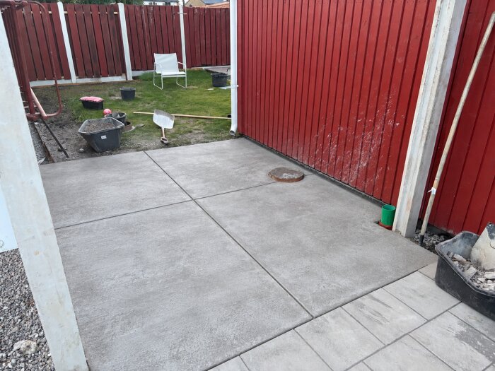Betongplatta i fyra sektioner framför ett rött plank, omgiven av trädgårdsredskap och en gräsmatta med en vit stol.
