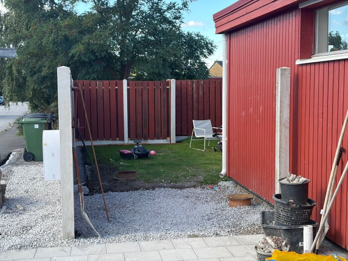 En trädgård under konstruktion med grusad yta, stolpar, röd träfasad och vit stol, leksaksbil och grön soptunna i bakgrunden.