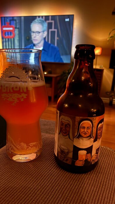Glas med öl och flaska märkt "Vreta Kloster Trippel" på ett bord, TV i bakgrunden som visar ett program.