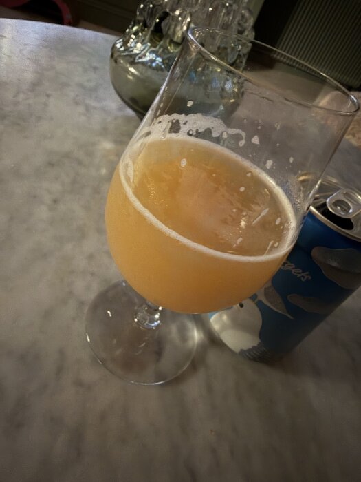 Ett glas med ljus, grumlig öl står på ett bord bredvid en öppnad ölburk med blå etikett.