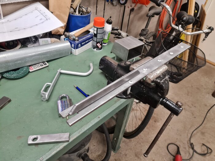 Verkstad med material för låsbom till garage, inklusive metallstång och skruvstycke, redo för svetsning.