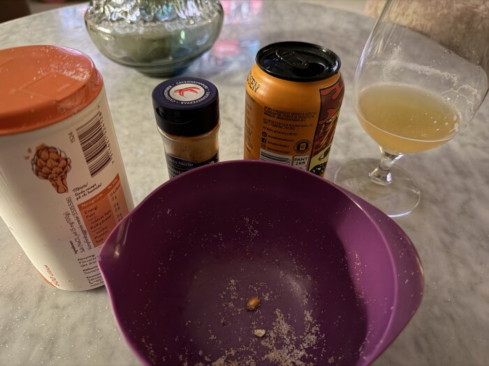 En skål med popcornrester, kryddburk, läskburk och glas med dryck på ett bord.