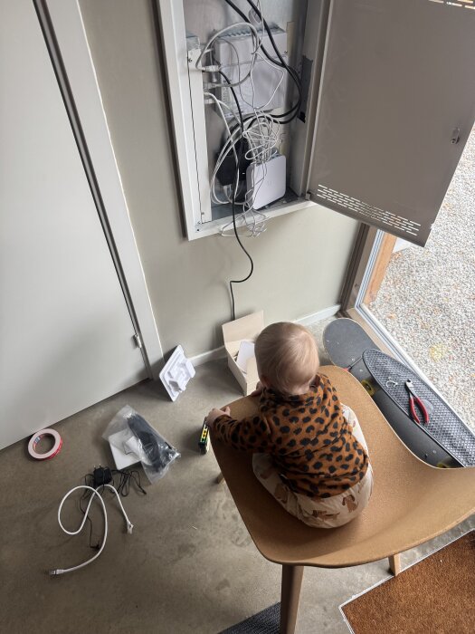 Barn i leopardmönstrad tröja sitter på en stol vid en öppen skåpdörr med nätverkskablar och utrustning, med verktyg och kablar på golvet.
