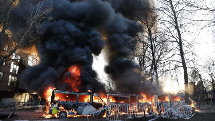 Brinnande bilar omgivna av tjock, svart rök i en stadsmiljö med träd och byggnader i bakgrunden.