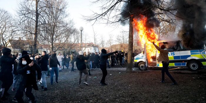 En grupp demonstranter samlade i en park, en brinnande polisbil, rök och lågor synliga.