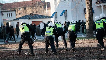 Poliser med hjälmar och skyddsvästar backar från en demonstration; en folkmassa syns i bakgrunden.