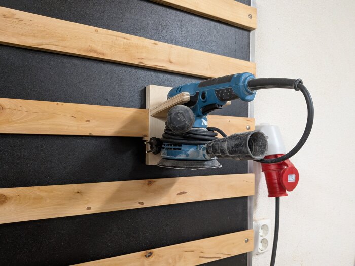 Blå slipmaskin monterad på vägg med träpanel, svart kabel hänger ner, eluttag syns i bakgrunden.