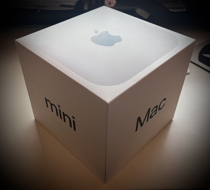 Vit kartong med texten "Mac mini" och Apple-logotyp på ovansidan, placerad på ett bord.