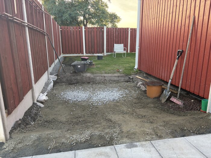 Utgrävning med uppfyllnad av bergkross och makadam vid ett rött plank och en husvägg, med verktyg och hinkar synliga.
