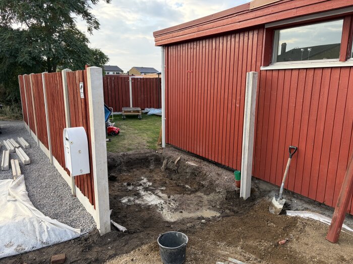 Utgrävning vid röd byggnad med röd träfasad, spade och skottkärra synliga, förbereder fyllning med bergkross och makadam.