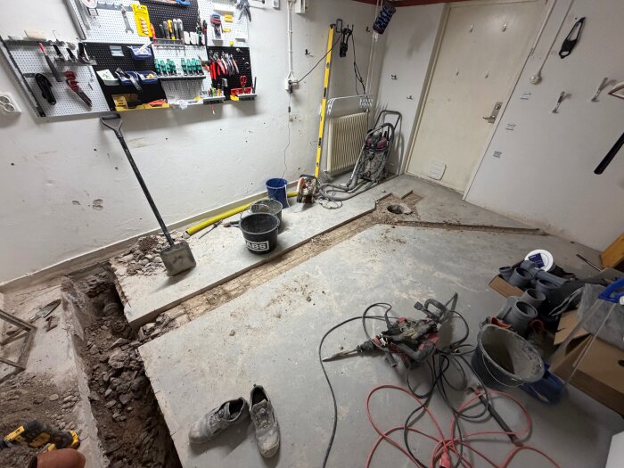 Källare under renovering med uppbilat golv för byte av avloppsrör. Verktyg, en spade och skyffel syns på golvet, samt ett verktygsstativ på väggen.