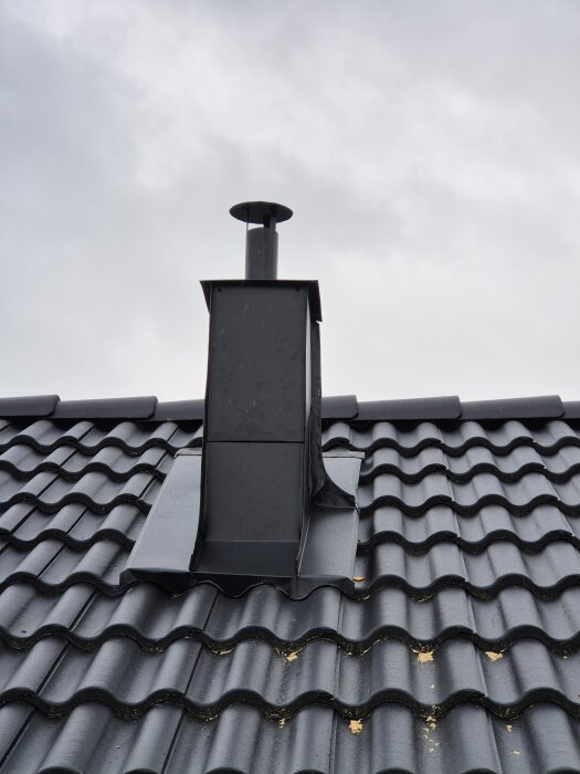 Ventilationshuv på tak, ser sned och knöglig ut, med svart plåt på svart tak med takpannor.