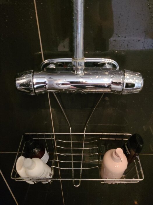 Duschkran med metallhylla innehållande schampoflaskor i ett mörkt kaklat badrum.