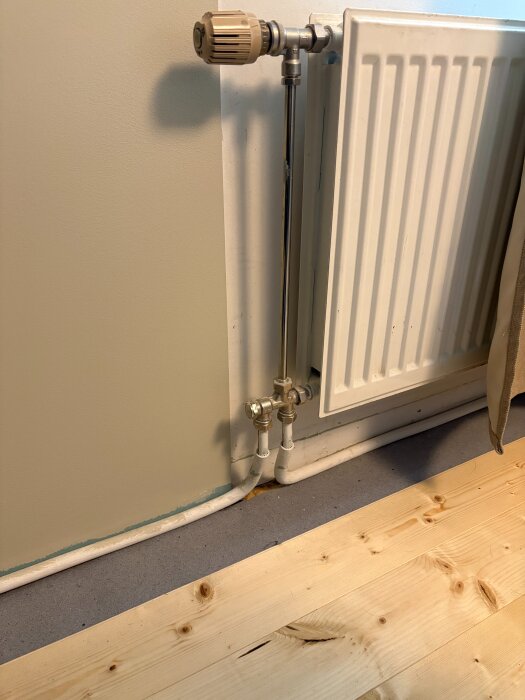 Radiator med kopparrör och termostatventil, installerad mot en vägg med parkettgolv.