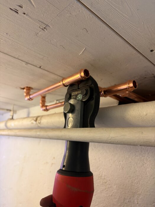 Pressverktyg används för att fästa kopparrör i ett värmesystem, synligt under taket med andra rör i bakgrunden.
