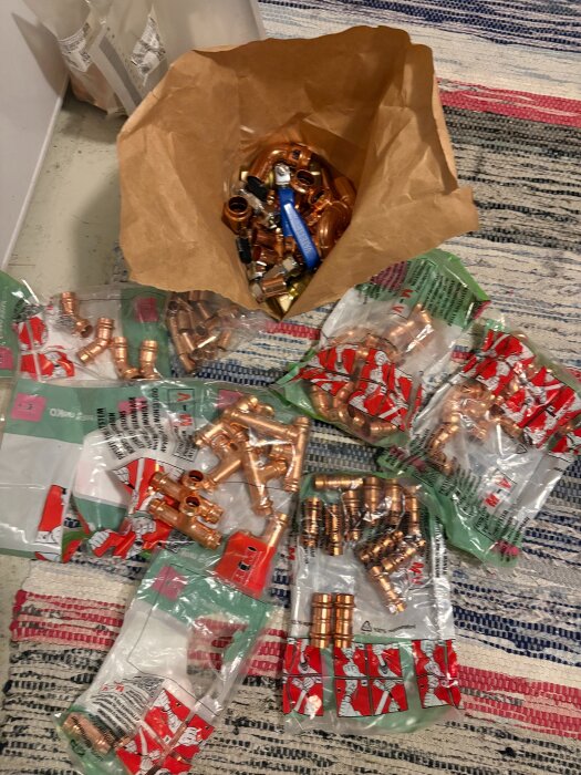 Kopparrör och rördelar i plastpåsar ligger på en matta, tillsammans med en brun påse fylld med fler kopparfittings och ett verktyg.