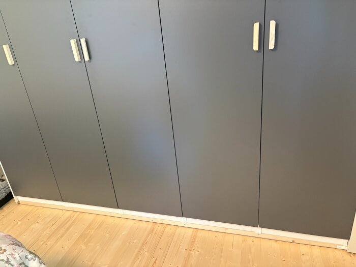 Gråa garderober med vita handtag, installerade på en låg sockel av reglar på ett trägolv i ett nästan färdigrenoverat sovrum.