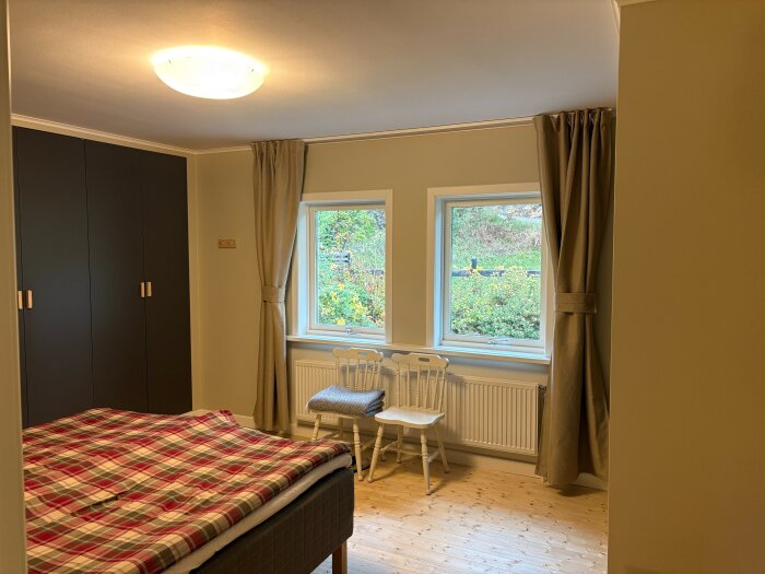 Sovrum med inbyggda mörka garderober, säng med röd-pläderad filt, två fönster med beige gardiner och två vita stolar framför ett element.