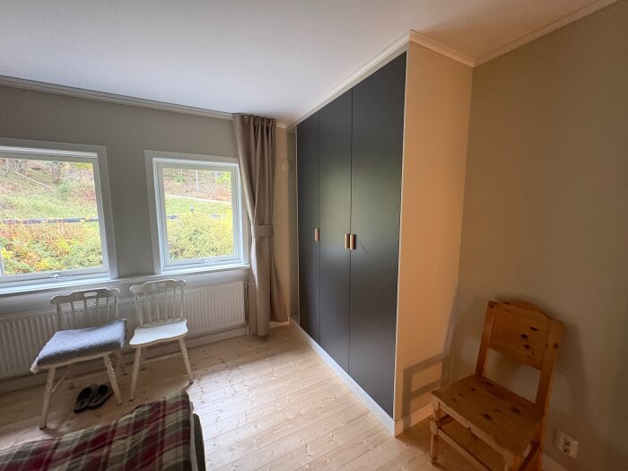 Sovrum med två fönster, mörka garderober inbyggda mot väggen och trästolar på ljust trägolv.