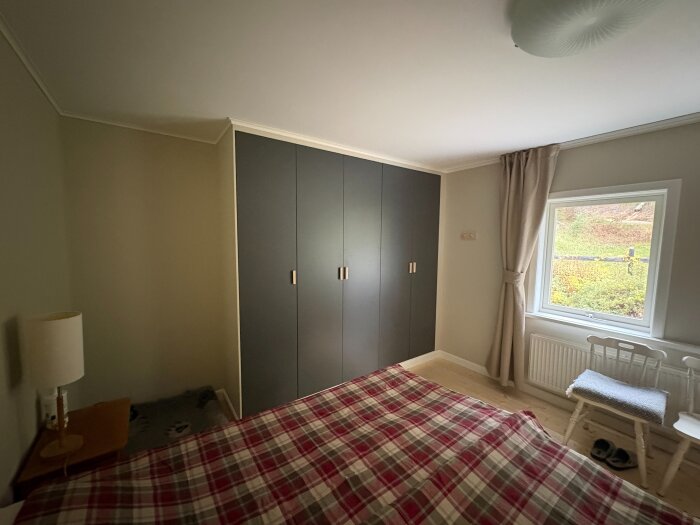 Sovrum med inbyggda garderober, fönster, och en säng med rutig överkast. Garderoberna står på en låg sockel.