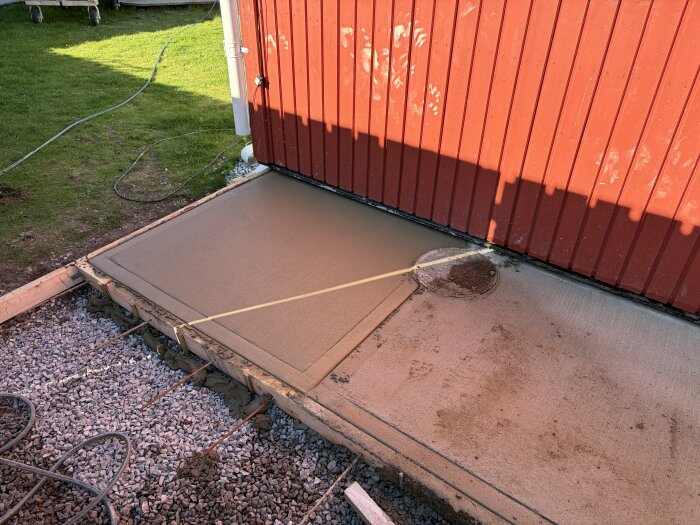 Nygjuten betongplatta framför rött trävägg. Området omges av grus och formar. En brunnslock syns på plattan. Solbelyst gräsmatta i bakgrunden.