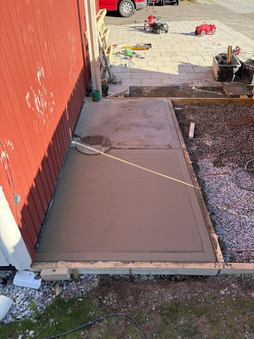 Nygjuten betongplatta vid en röd byggnad, omgiven av stenläggning och grus. Leksaksbilar och byggverktyg ligger på marken.