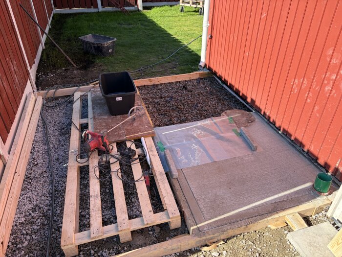 Byggarbetsplats för betongplatta vid röd byggnad, med träpallar, verktyg och elektriska kablar på marken.