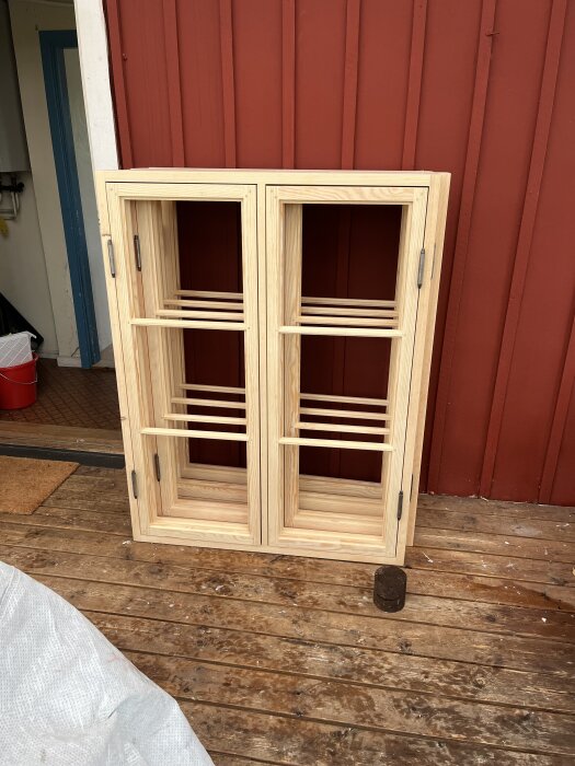Nya omålade fönsterbågar står mot en röd trävägg på en altan, redo för glasning och målning.