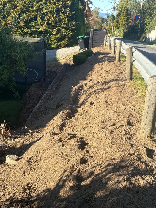 Brant slänt utan planter, täckt med jord, redo för plantering av thuja. Stenblock och stängsel vid sidan.
