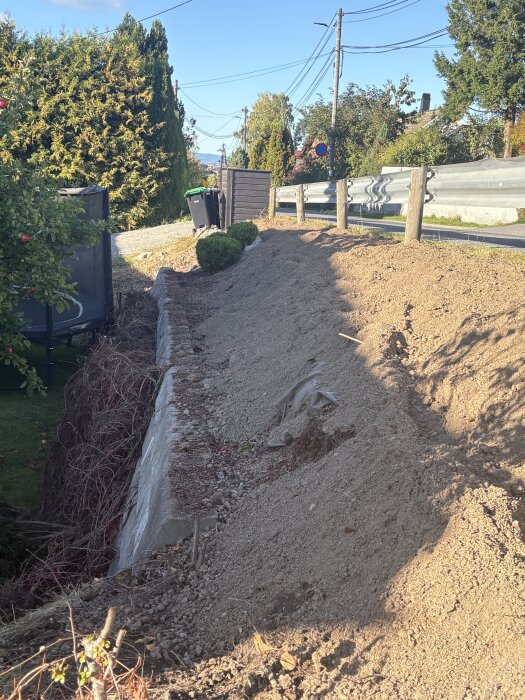 Brant slänt med nylagd planteringsjord, omgiven av växter och staket, sedd vid en väg. Förberedelse för plantering och erosionstabilisering.