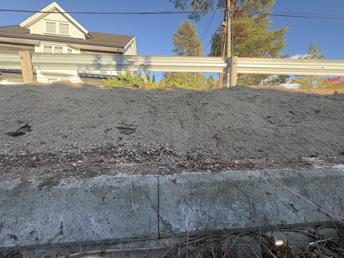 En hög med grus framför ett stålräcke, med ett stort hus och skog i bakgrunden under en klarblå himmel.