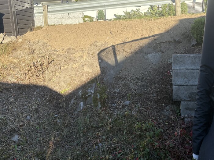 En jordhög mot en stödmur, omgiven av växtlighet och en skugga från ett staket i närheten.