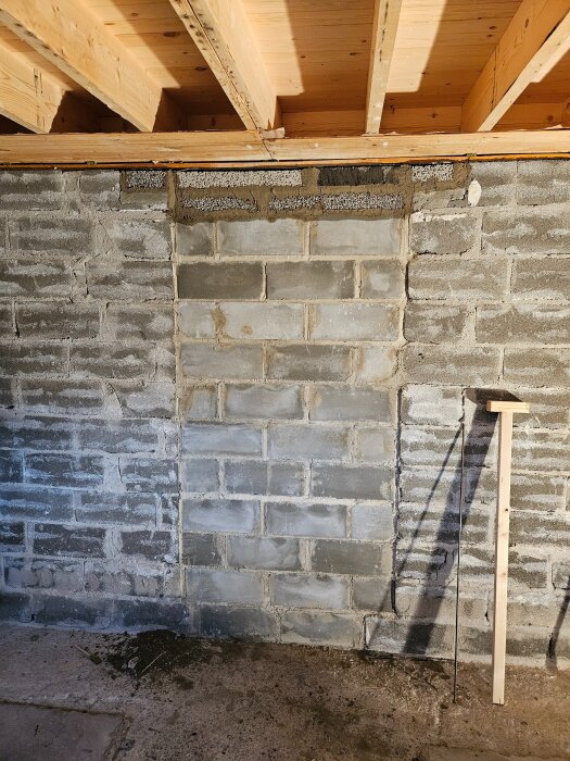 En vägg av grå betongblock i en källare, med ett träbjälklag ovanför. En mätsticka står lutad mot väggen.