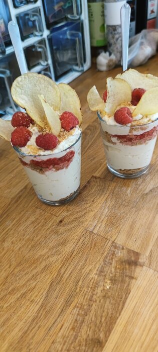 Glas med cheesecake toppade med hallon och chips på ett träbord, några ingredienser syns i bakgrunden.