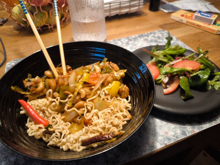 Bilden visar en skål med thai kyckling med cashewnötter och chili på nudlar, med en sallad på en tallrik bredvid.