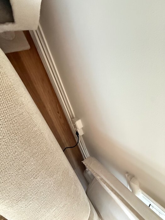 Vägguttag med en kabel ansluten, beläget på en vit vägg bakom en beige soffa i ett vardagsrum.