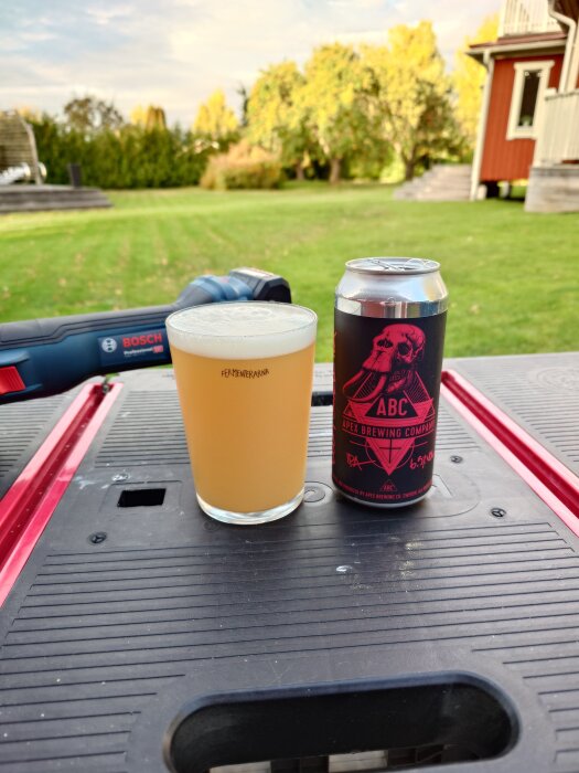 Ölglas och burk med Apex - Doom Swell IPA från Apex Brewing Company på ett bord i en trädgård, med träd och hus i bakgrunden.