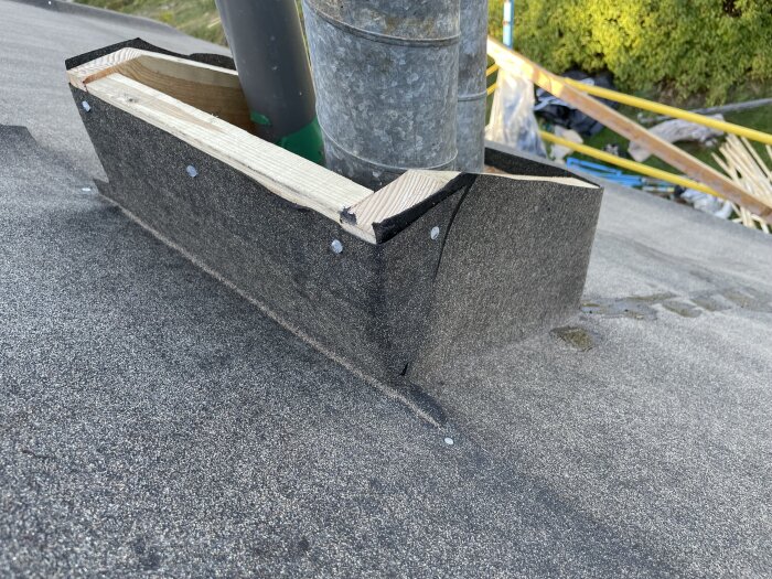 Träram av 30 mm virke runt ventilationsrör, täckt med tjockt material för genomföring på tak. Spikar och sömmar synliga vid kanterna.