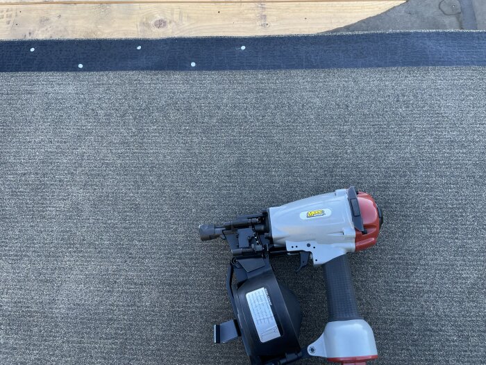 Spikpistol på grå taktäckning med synliga skruvar längs en svart remsa, träyta i bakgrunden.
