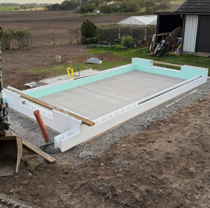 Poolbygge i trädgården med thermoblock placerade för väggar, omgiven av jord och betongplatta i botten. Trädgård och verktyg syns i bakgrunden.