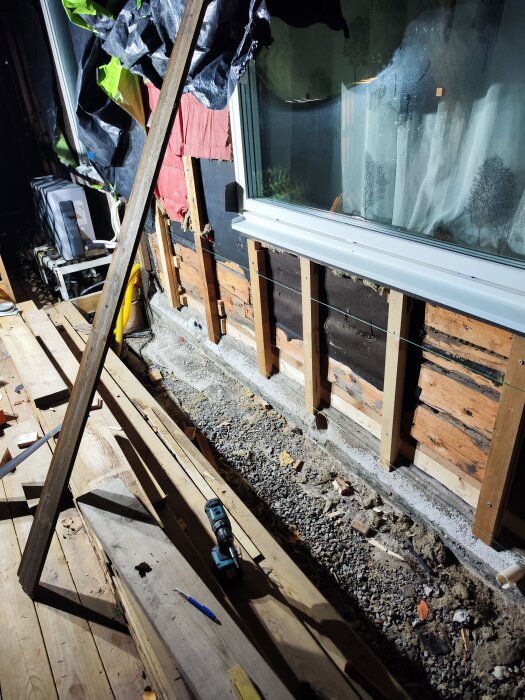 Vägg under renovering med exponerade träreglar, fönster och verktyg på marken, diskuteras byte av tjärpapp och isolering.