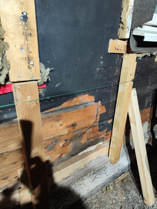 Vägg med träreglar, exponerade bjälkar och sliten tjärpapp i ett byggprojekt.