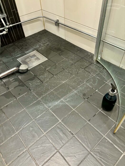 Grått klinkergolv i en duschkabin med vita kalkfläckar på ytan, rörledningar längs väggen och duschhuvud på golvet.