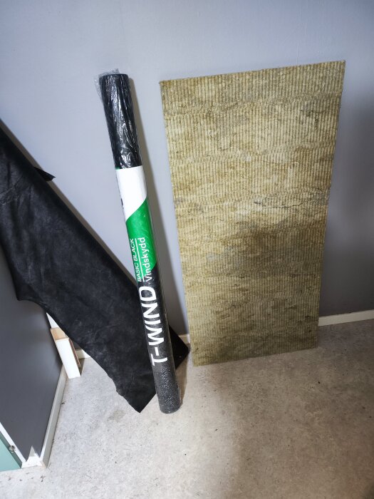 Rulle med vindskydd, svart underlagspapp och skiva av isoleringsmaterial placerade mot en vägg.