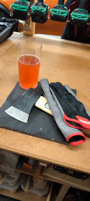 En yxa, arbetshandskar och glas med orange dryck på träarbetsbänk i verkstadsmiljö.