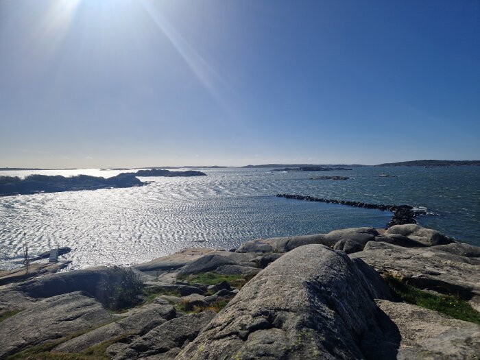 Klippor vid soligt kustlandskap med havsutsikt och vågor som sköljer mot stenarna.