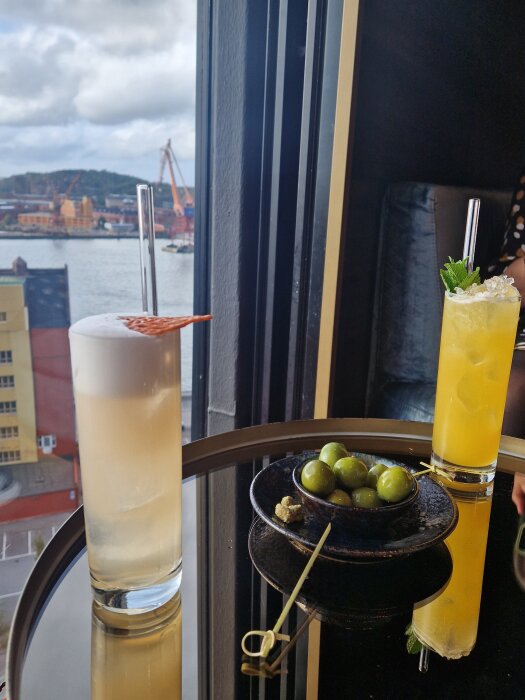 Två cocktails på ett bord med utsikt över hamn; ett glas med skum och en med mynta. En skål med gröna oliver mellan glasen.