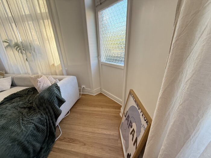 Soffa med kuddar och filt invid ett fönster, elkabel synlig längs golvet, en tavla står lutad mot väggen nära balkongdörren.
