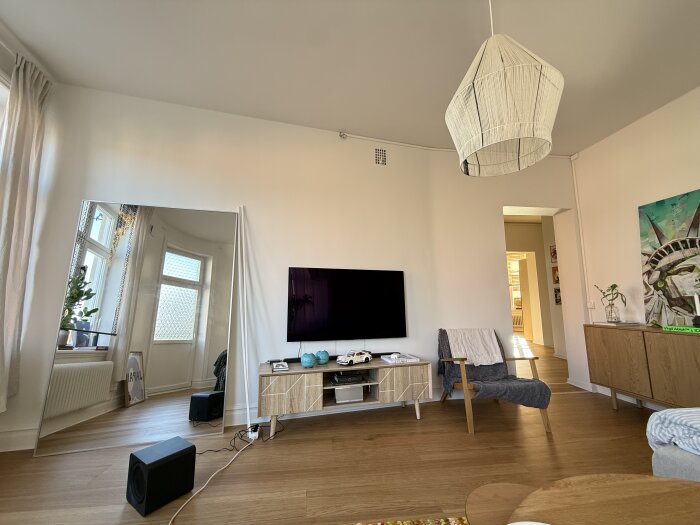 Modernt vardagsrum med TV på väggen, stor spegel, fåtölj och trägolv. Synliga kablar på golvet anslutna till en subwoofer.